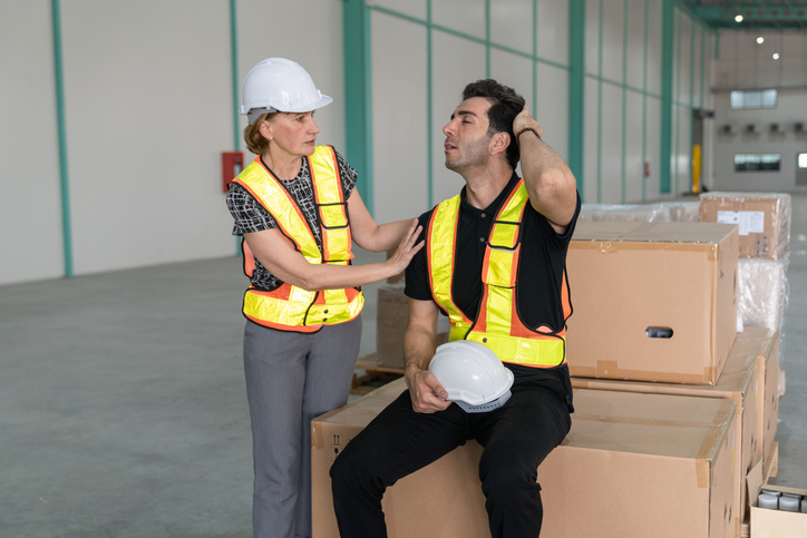 Woman factory engineer comforts a strained male engineer inside a large industrial warehouse.Male factory engineer with head injury sitting on cargo box with female engineer helping.