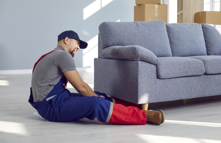 Man gets seriously injured while trying to lift heavy furniture alone. Professional handyman who got his foot caught under sofa sitting on floor and crying. Work injury, broken foot, pain concept